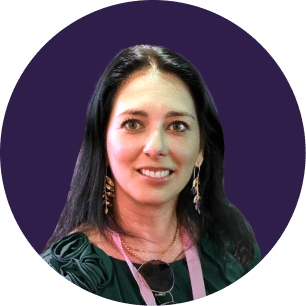 Mujer con el pelo largo y oscuro, que lleva pendientes de oro y un top verde, y sonríe a la cámara. Lleva un cordón colgado del cuello. El fondo es un círculo liso de color morado oscuro.