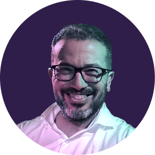 Hombre sonriente con gafas y barba, vestido con una camisa blanca, sobre un fondo circular liso de color morado.