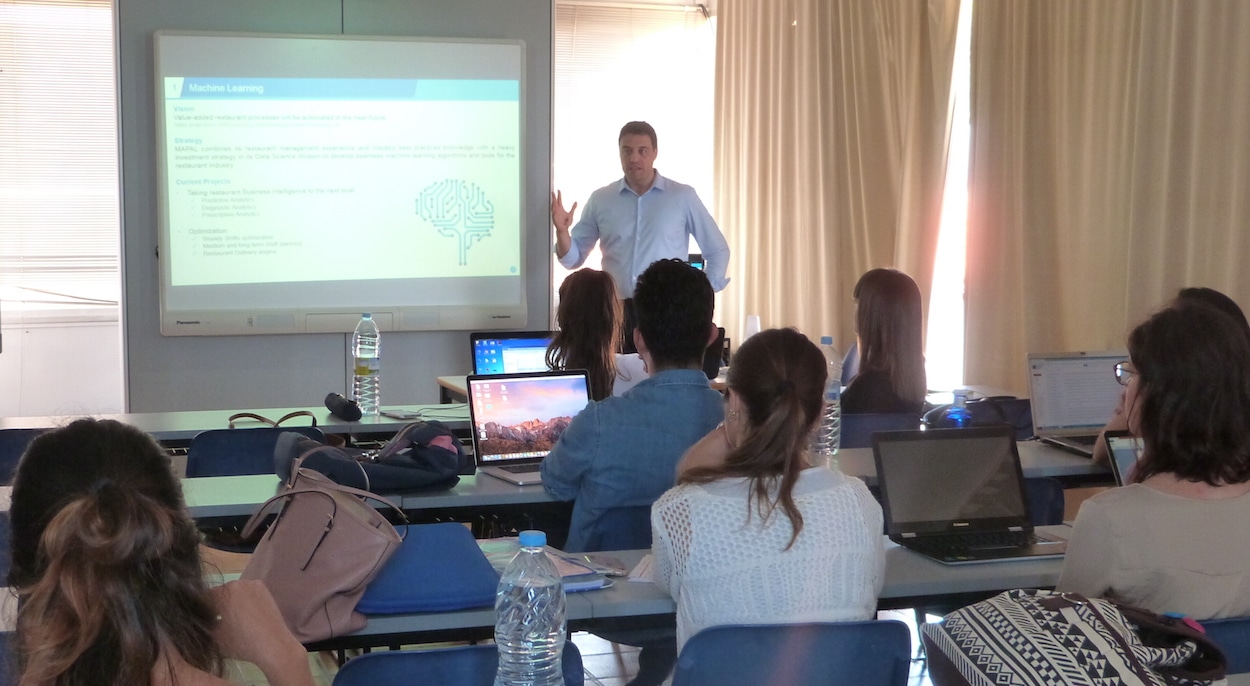 Un hombre está de pie frente a un aula impartiendo una presentación sobre aprendizaje automático. Varios alumnos están sentados en las mesas con sus ordenadores portátiles abiertos, escuchando y tomando apuntes. En la pantalla se ve una diapositiva proyectada con texto y un gráfico de un cerebro.