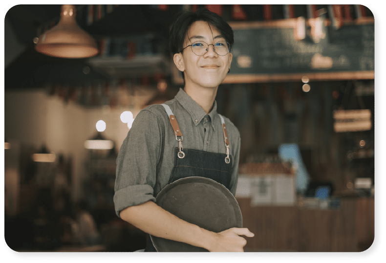 Una persona con gafas, una camiseta gris y un delantal oscuro sonríe mientras sostiene una bandeja redonda en una cafetería con una iluminación cálida, con luces y elementos decorativos difuminados al fondo.