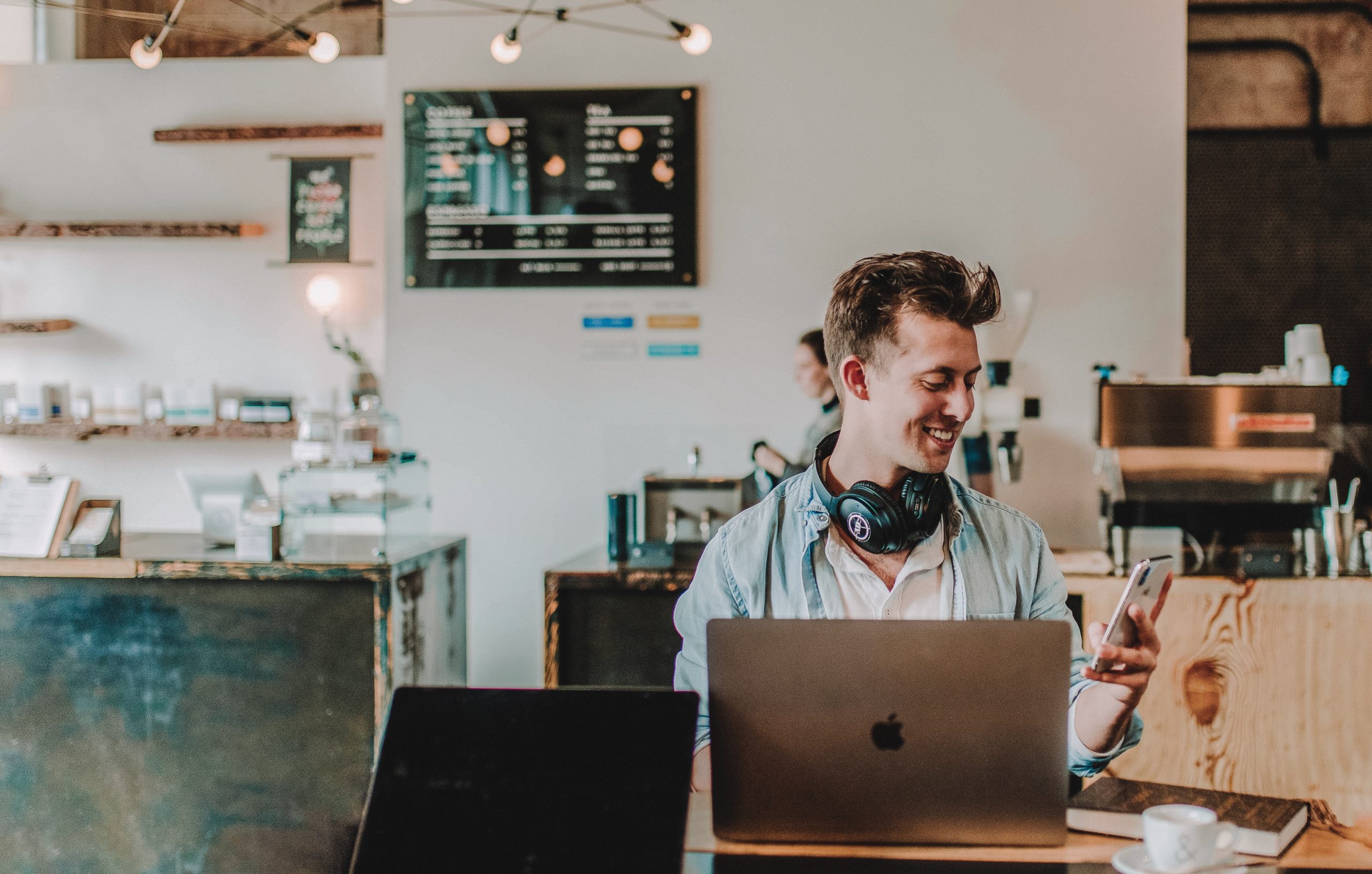 Una persona sonriente está sentada en la mesa de una cafetería con un ordenador portátil, un smartphone y unos auriculares colgados del cuello, rodeada por la decoración y el equipamiento de la cafetería.