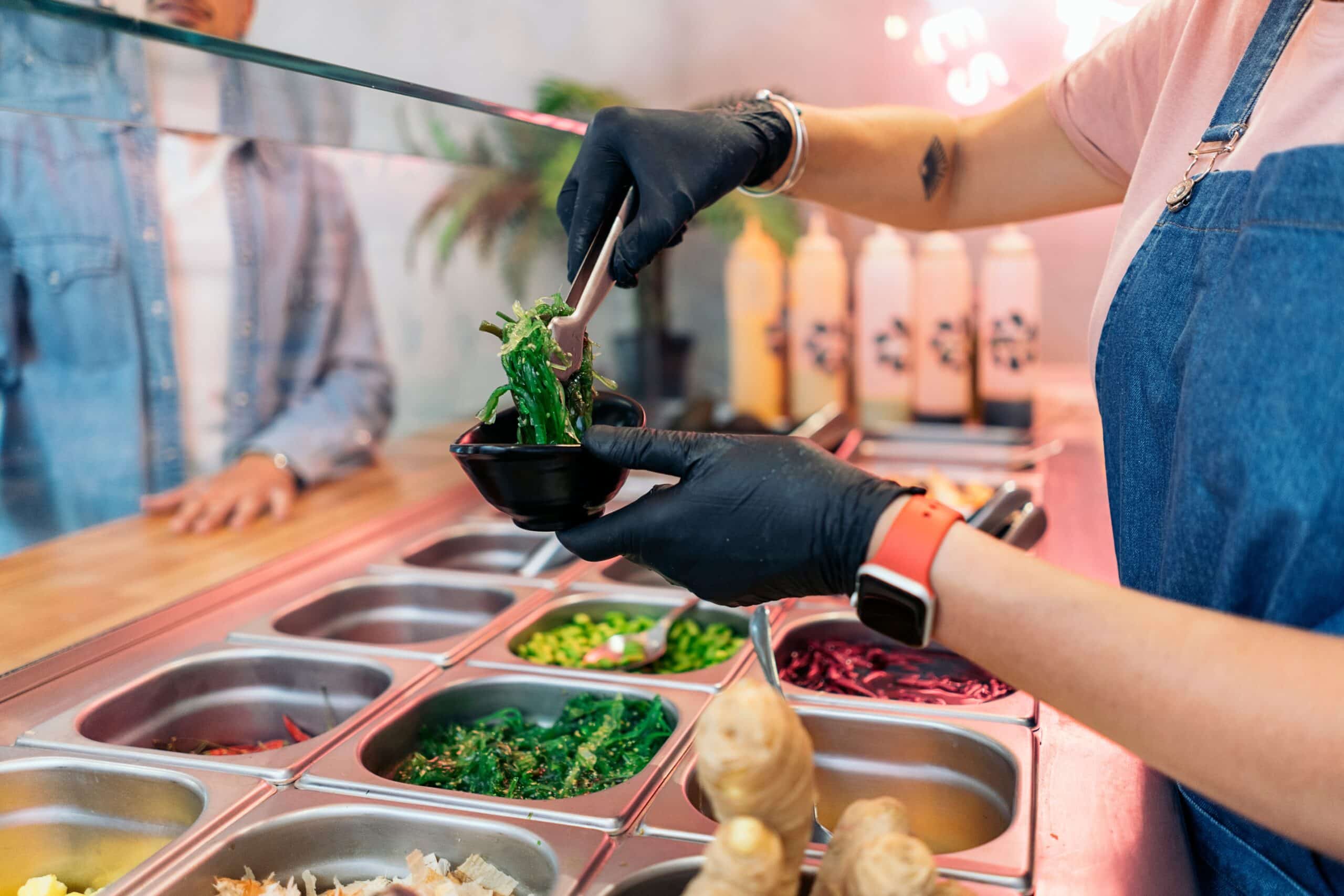 Una persona que lleva guantes y delantal sirve lechuga en un cuenco pequeño en una barra de ensaladas, donde hay diversos ingredientes frescos en recipientes metálicos.