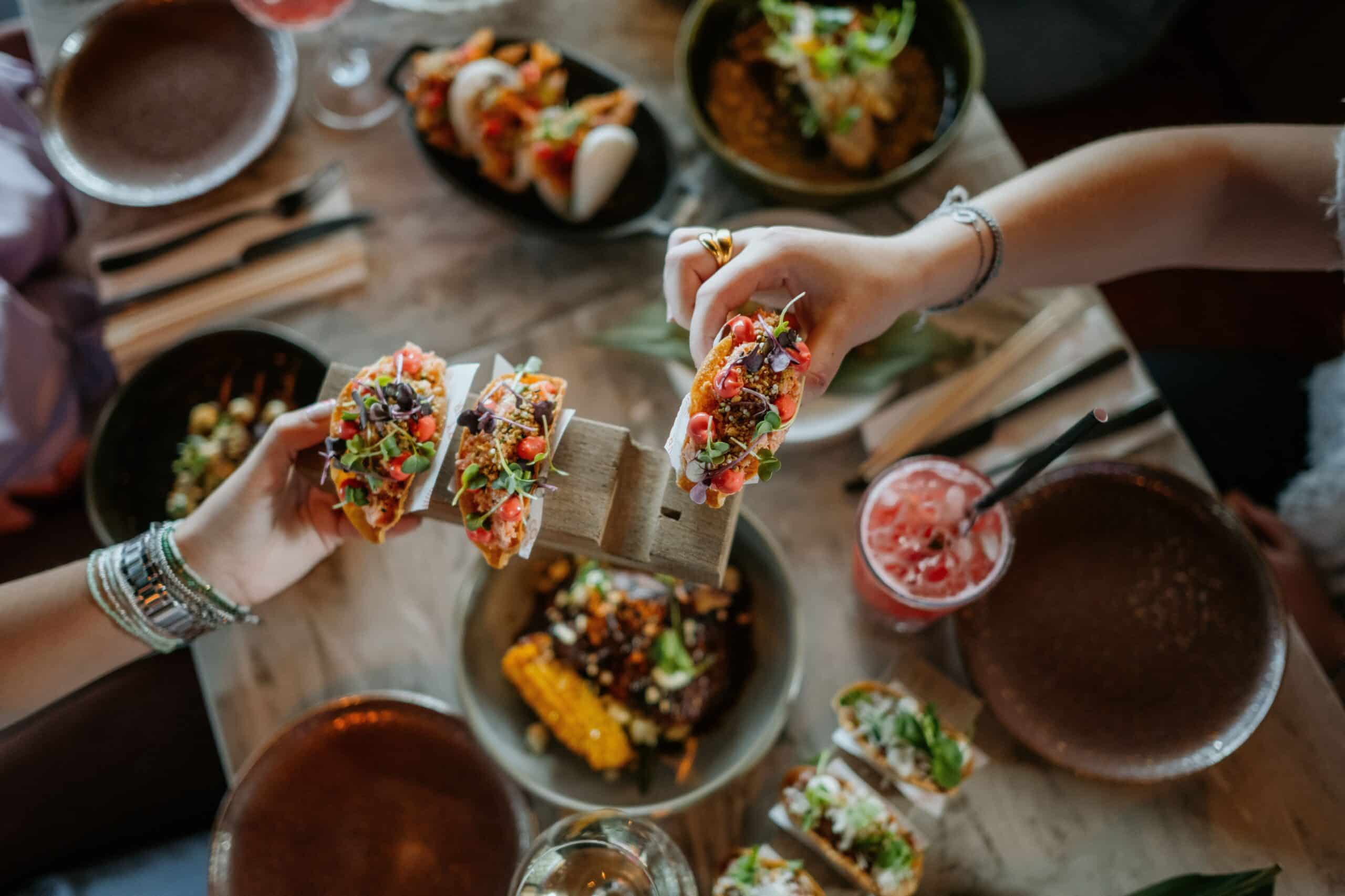 Dos personas sostienen unos tacos de colores sobre una mesa repleta de platos diversos, entre los que se incluyen mazorcas de maíz, panecillos bao y bebidas rosas, lo que sugiere una animada reunión para comer.
