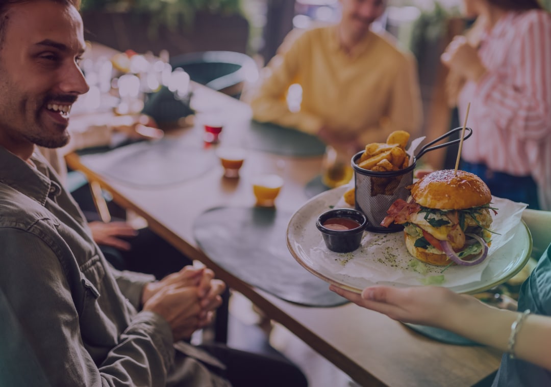 Una persona sentada en la mesa de un restaurante sonríe mientras alguien le sirve un plato con una hamburguesa, patatas fritas en un vaso de metal y un cuenco pequeño con salsa. Al fondo hay otras personas sentadas, disfrutando de la comida juntos.