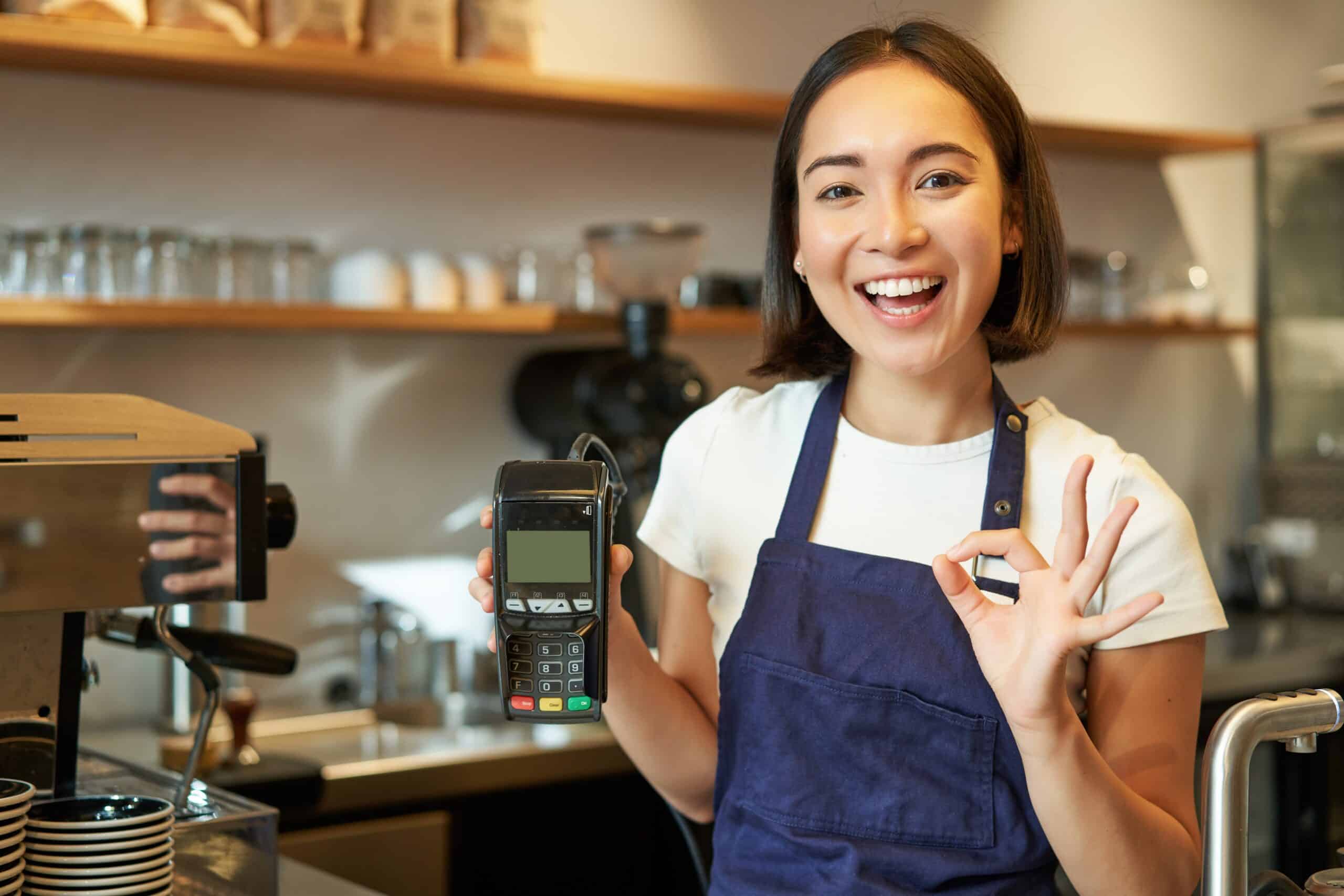 Un barista sonriente, con un delantal azul, sostiene un terminal de pago en una mano y hace un gesto de «OK» con la otra, de pie detrás del mostrador de una cafetería.
