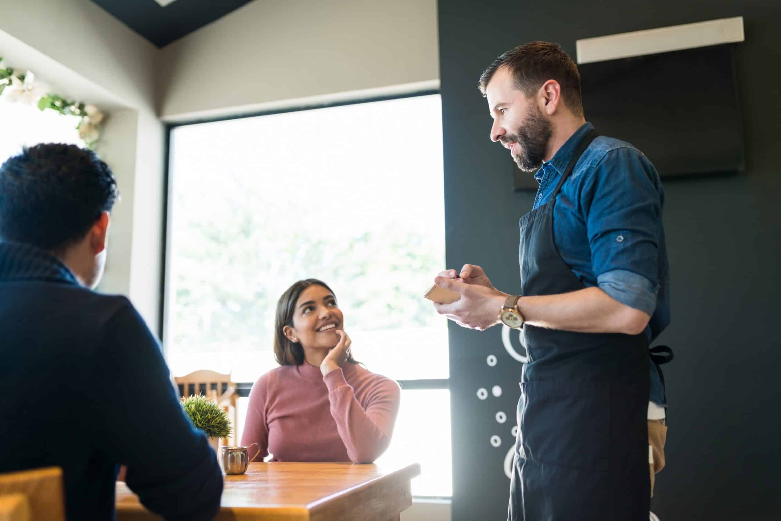 Un camarero sonriente, con delantal, habla con dos personas sentadas en una mesa de restaurante junto a una ventana soleada. Una de ellas levanta la vista, escucha y sonríe, mientras que la otra está sentada de espaldas a la cámara.