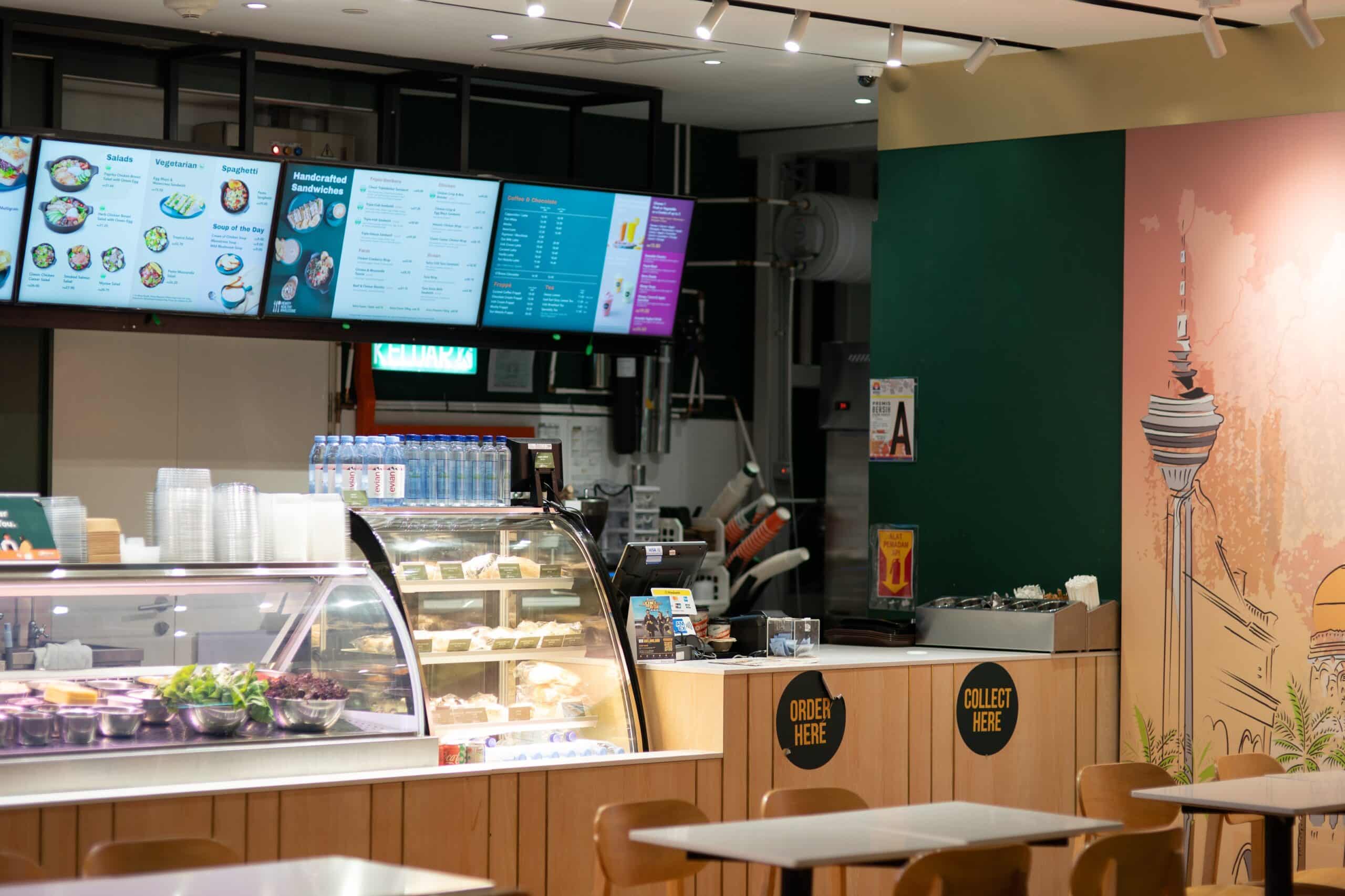 En la barra de una cafetería, muy bien iluminada, se exponen ensaladas y bebidas, con pantallas digitales con el menú colocadas encima. Los carteles indican «Pedir aquí» y «Recoger aquí». En primer plano hay mesas y sillas; un mural decora la pared.
