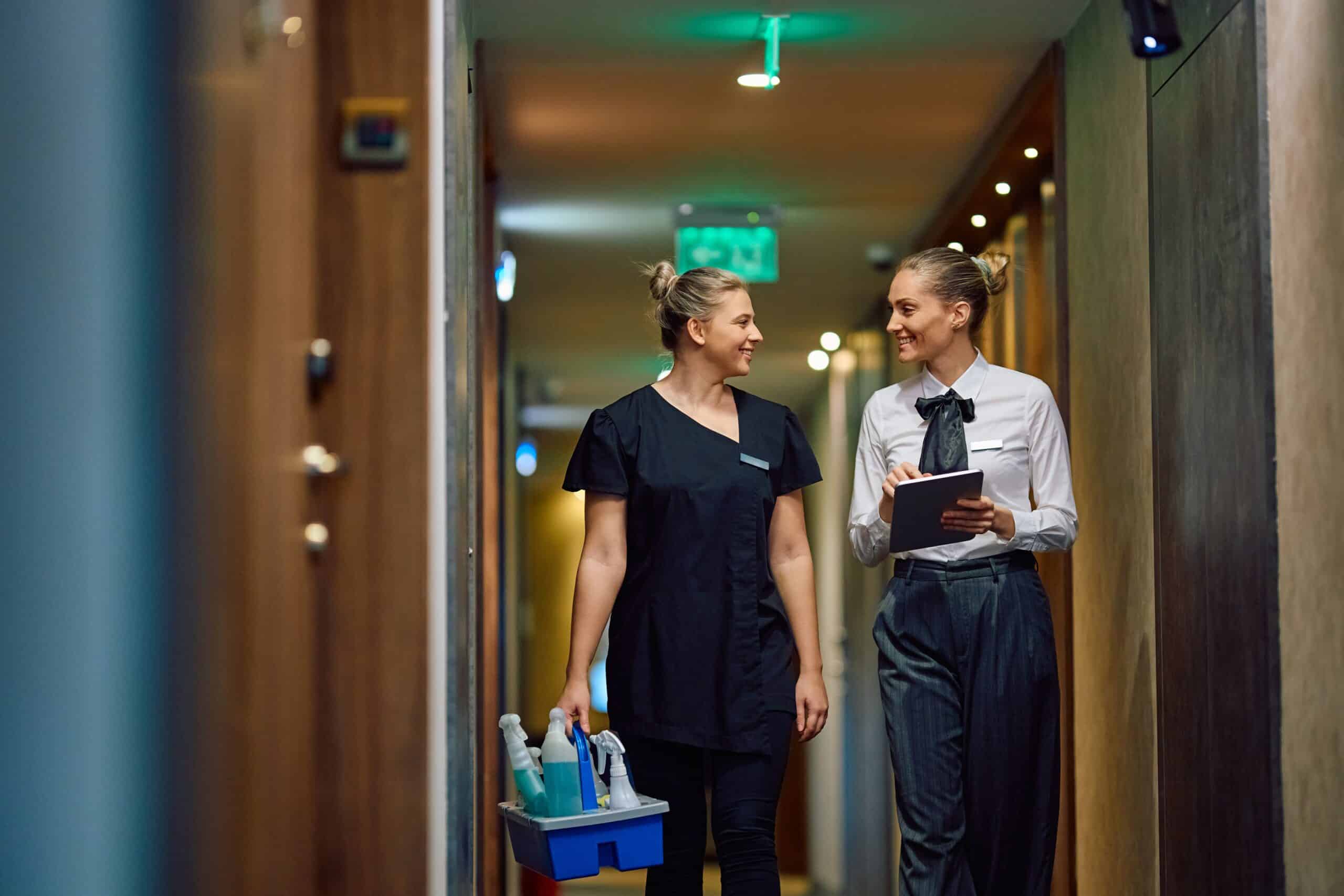 Dos mujeres caminan por un pasillo: una lleva un uniforme de limpieza y transporta productos de limpieza, y la otra, vestida con traje de negocios, sostiene una tableta. Ambas sonríen y parecen estar conversando.