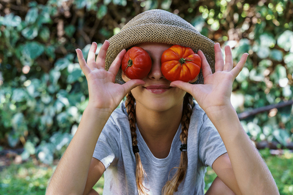 Un niño con un sombrero de paja y una camiseta gris se tapa los ojos con dos tomates, sonriendo juguetonamente al aire libre con vegetación de fondo.