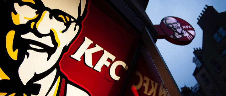 Primer plano del letrero de un restaurante KFC con el logotipo del Coronel Sanders, en el que se aprecia otro letrero circular de KFC al fondo, con el cielo del atardecer y las siluetas de los edificios como telón de fondo.