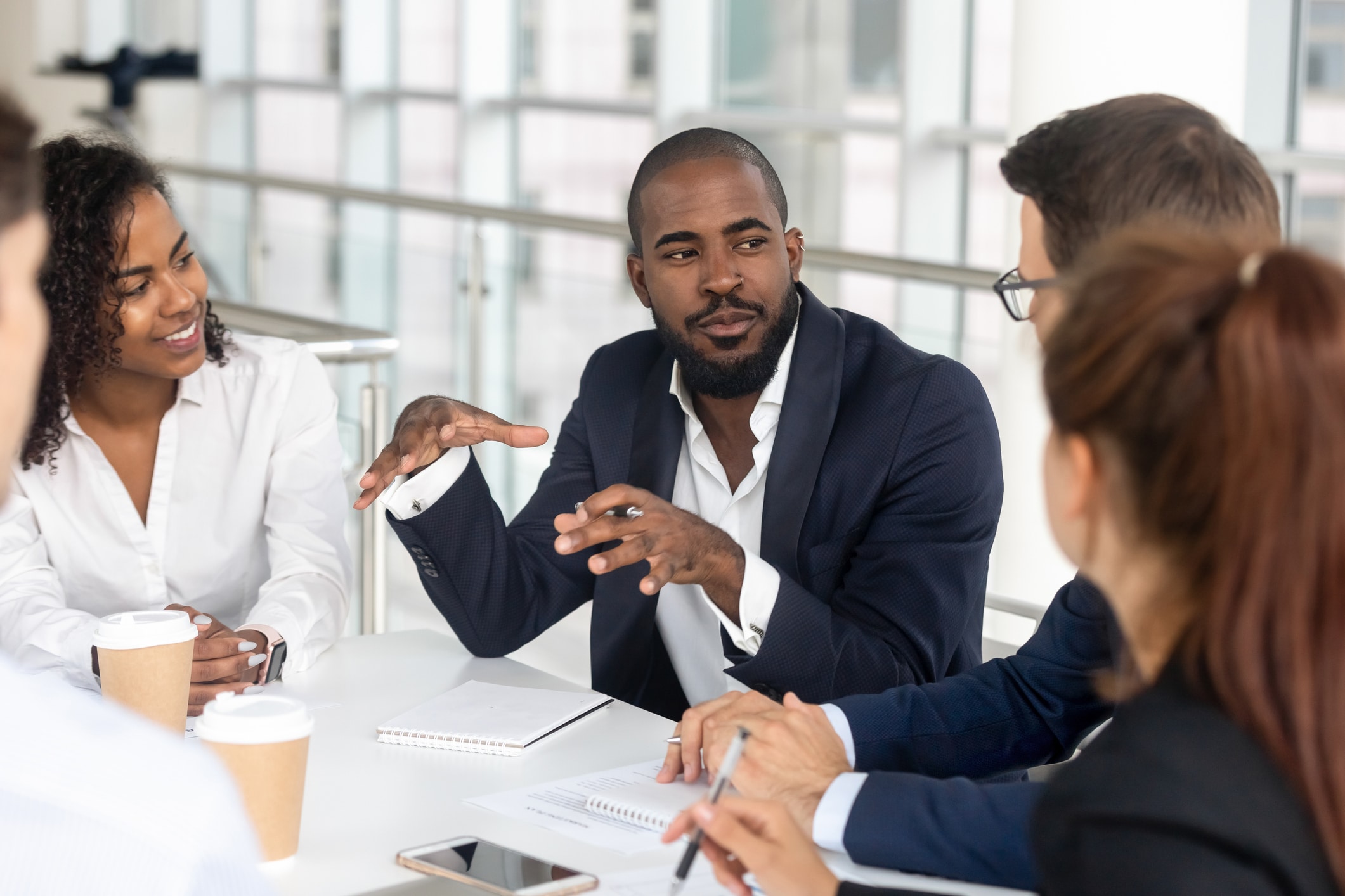 Cuatro personas vestidas con ropa de trabajo están sentadas alrededor de una mesa con cuadernos y tazas de café, inmersas en una conversación. Un hombre gesticula mientras habla y los demás escuchan con atención. Un entorno de oficina luminoso con grandes ventanas.