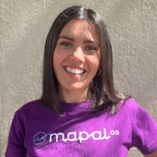 Una mujer de pelo liso y oscuro sonríe a la cámara; lleva una camiseta morada con el logotipo de Mapal Os. Está de pie frente a una pared beige con textura, bajo una luz natural muy intensa.