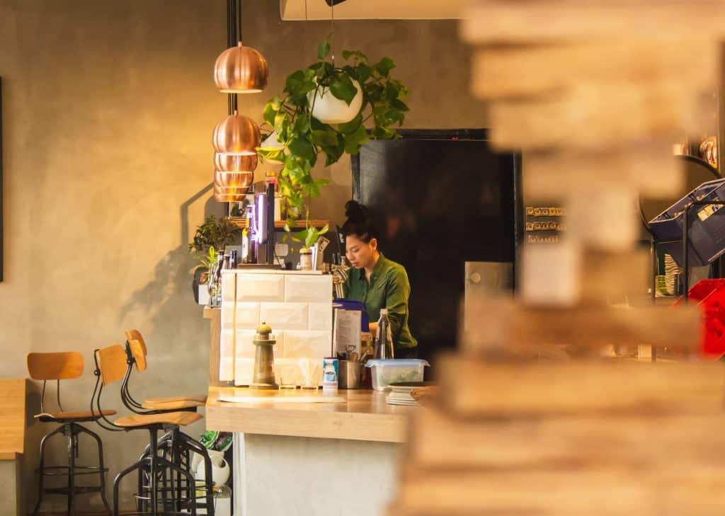 Una mujer está de pie detrás de la barra de una cafetería, hablando por teléfono. La cafetería tiene sillas de madera, lámparas colgantes, plantas y una iluminación cálida. El primer plano está desenfocado, y la atención se centra en la mujer que está trabajando.