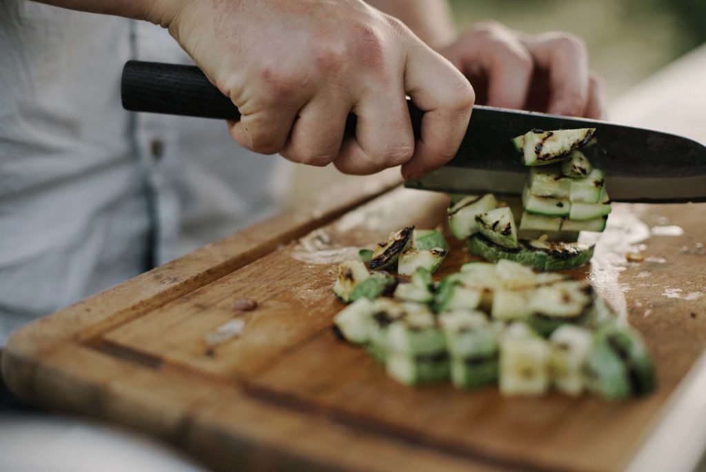 Primer plano de unas manos cortando calabacín a la parrilla sobre una tabla de cortar de madera con un cuchillo de mango negro.