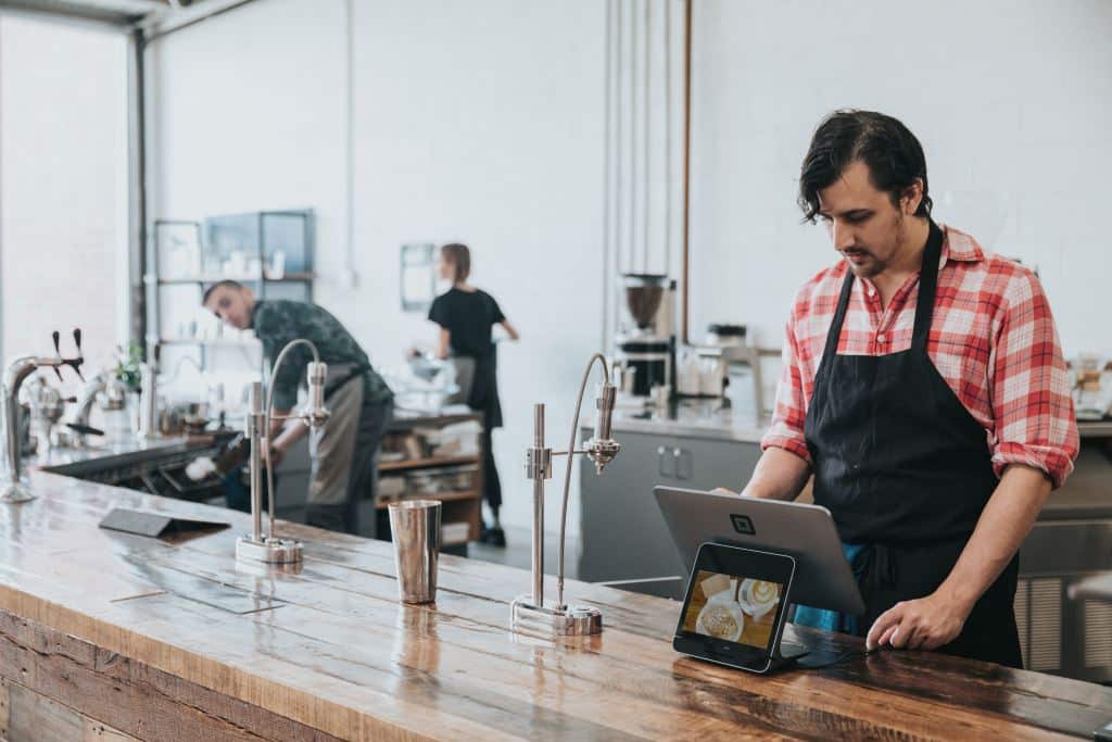 Un barista, con una camisa a cuadros rojos y un delantal negro, utiliza una tableta en la barra de una cafetería, mientras otras dos personas trabajan al fondo, cerca de la maquinaria de café y las estanterías.
