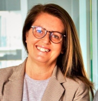 Una mujer con el pelo largo y castaño y gafas sonríe vestida con una camisa gris claro y una chaqueta de cuadros, de pie en el interior, frente a unos paneles de cristal.