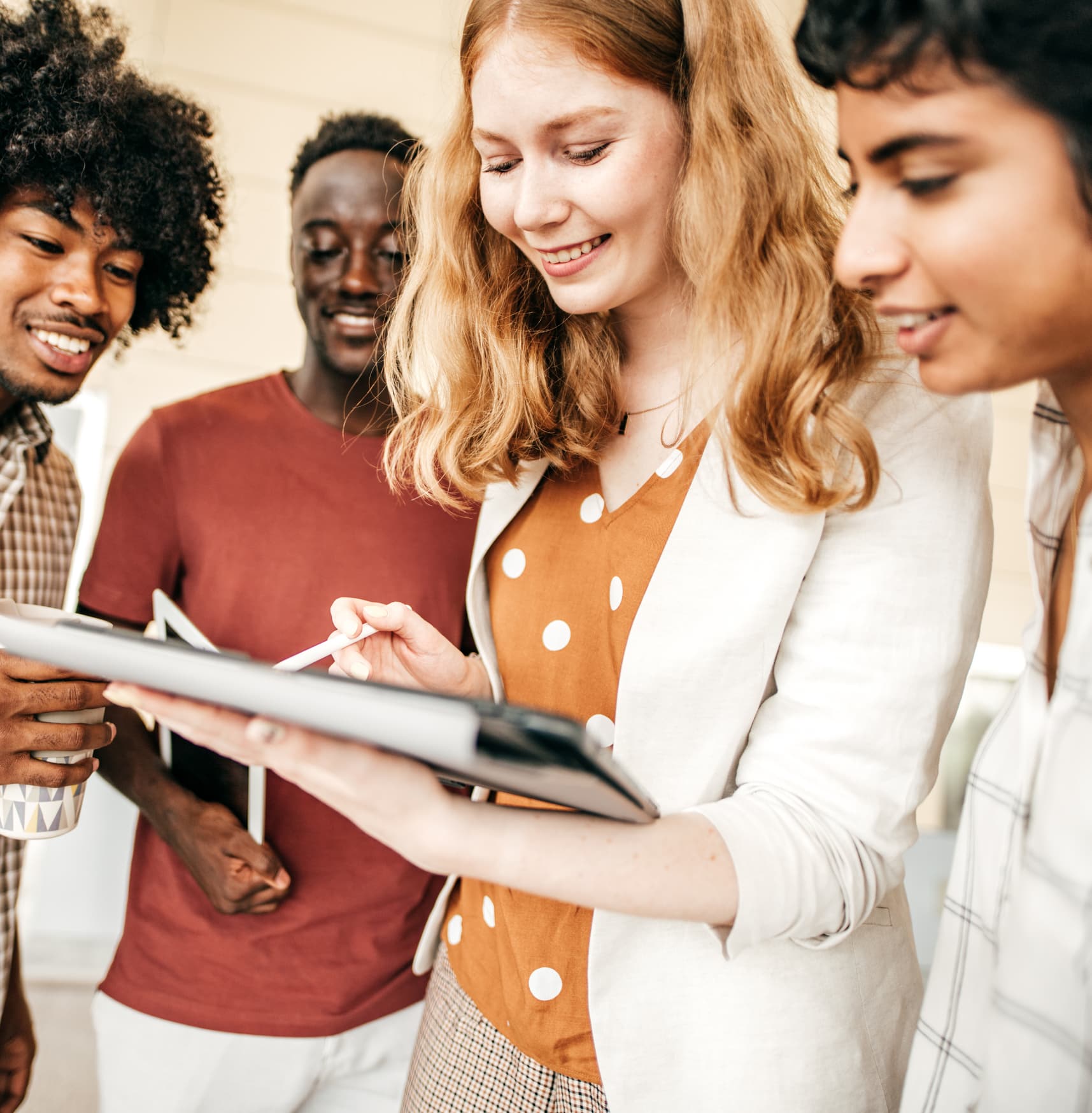 Cuatro jóvenes están muy juntos, sonriendo y mirando una tableta digital que uno de ellos sostiene y maneja con un lápiz óptico. Parece que están colaborando o trabajando en un proyecto.
