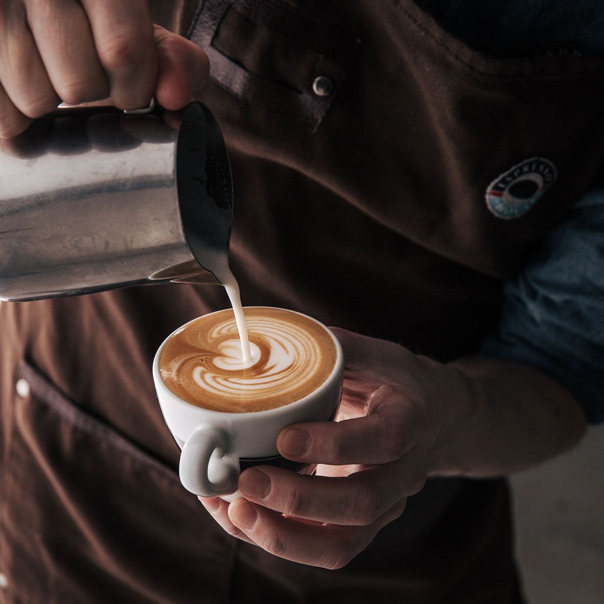 Uma pessoa que usa um avental castanho deita leite vaporizado de um jarro de metal numa chávena de café, criando uma figura na espuma.