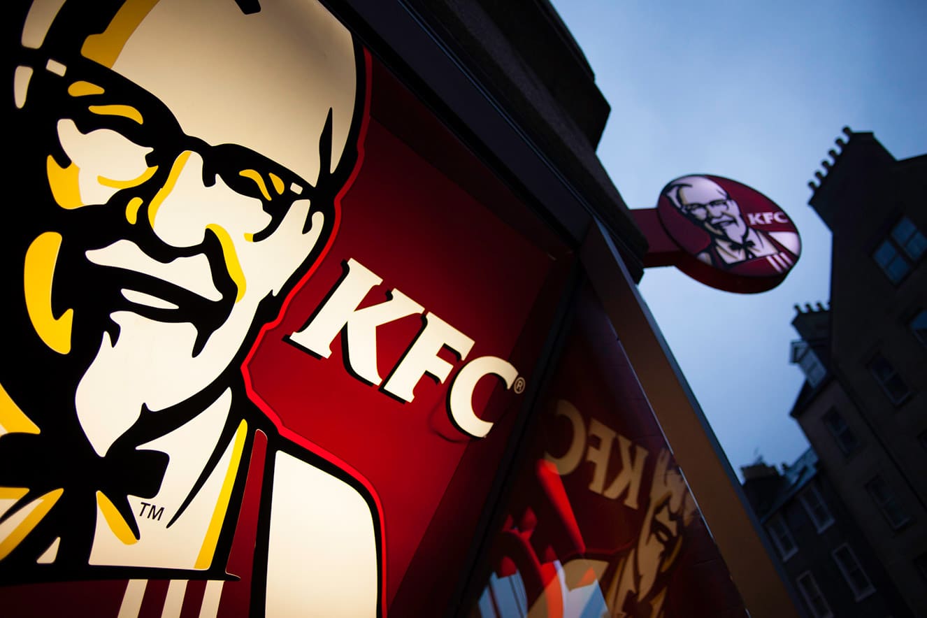 Grande plano da fachada de uma loja do KFC, com o logótipo do Coronel Sanders e o nome KFC, sendo visível um letreiro exterior do KFC contra um céu que escurece ao anoitecer.