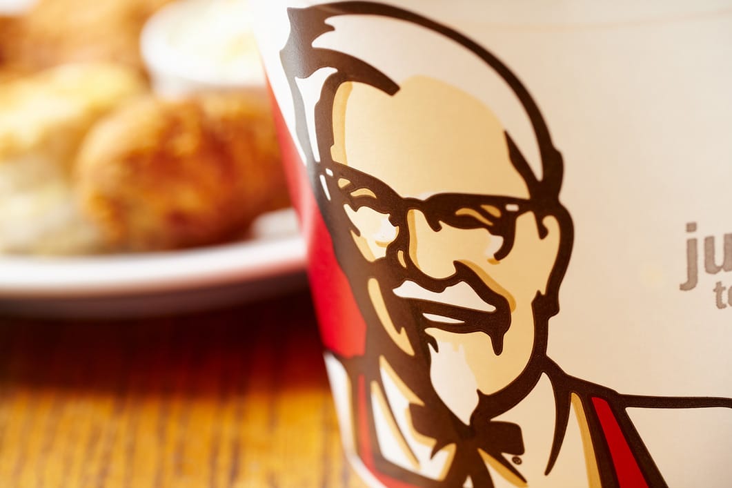 Grande plano de um balde do KFC com o rosto e o logótipo do Coronel Sanders, com frango frito e bolinhos difusos ao fundo, sobre uma mesa de madeira.