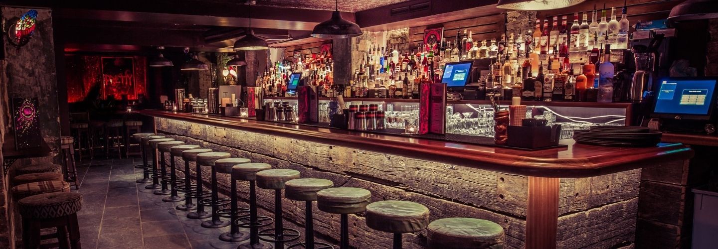 Um bar com pouca luz, com um longo balcão de madeira, bancos de bar, prateleiras cheias de garrafas de bebidas alcoólicas, copos e luzes suspensas brilhantes no teto.