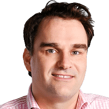 A man with fair skin, short dark brown hair, and blue eyes, wearing a light pink collared shirt, smiles slightly against a plain white background.