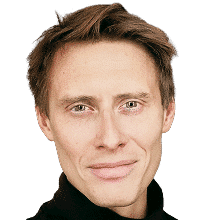 A man with light skin, short brown hair, and blue eyes is facing the camera and smiling slightly. He is wearing a black shirt and is set against a plain white background.