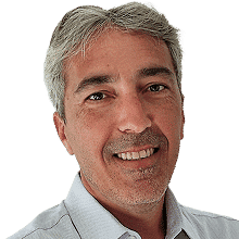 A middle-aged man with short grey hair, wearing a light-coloured collared shirt, smiling at the camera against a plain white background.