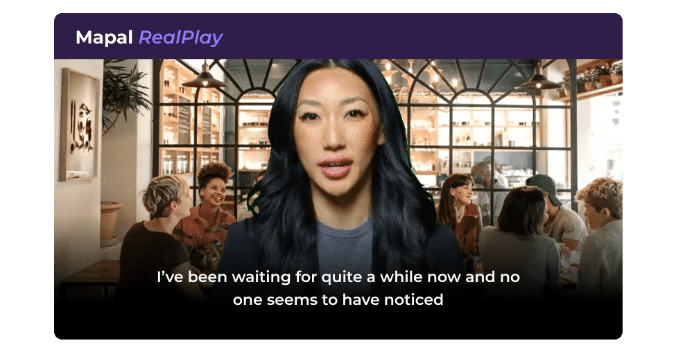 A woman with long black hair speaks in front of a blurred background of people sitting and talking in a café. The subtitle reads: I've been waiting for quite a while now and no one seems to have noticed.