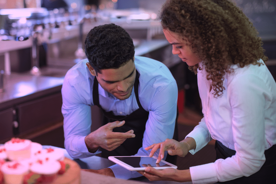 Two people in work uniforms look at a tablet together in a kitchen. One person is pointing at the screen whilst the other touches it. Cupcakes are visible in the foreground.