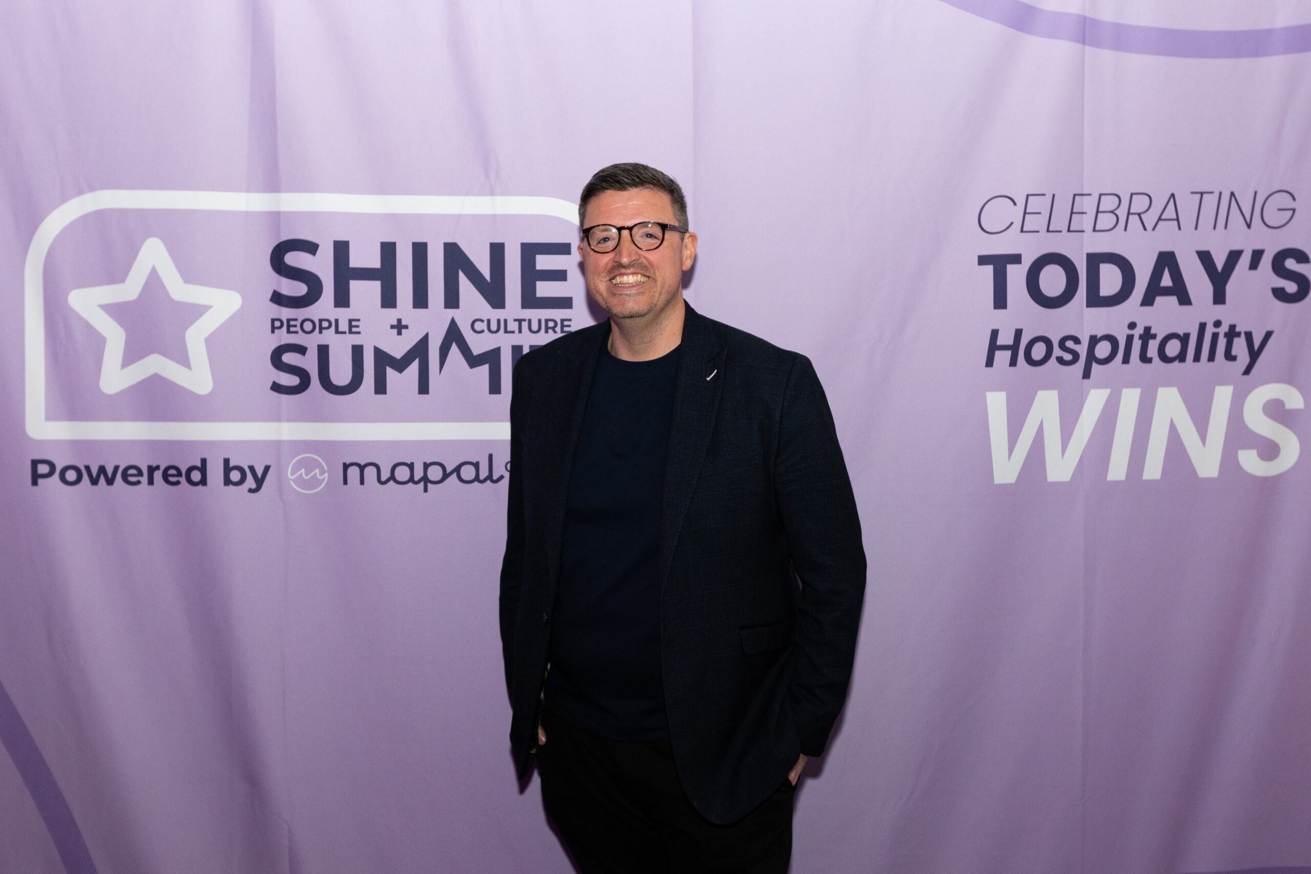 A man in glasses and a dark suit stands smiling in front of a purple backdrop with Shine Summit and Celebrating Today's Hospitality Wins written on it.