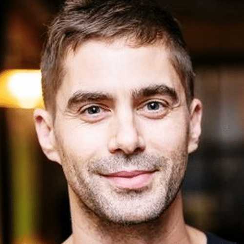 A man with short brown hair and a trimmed beard smiles gently at the camera, standing indoors with a blurred background that includes a warm light.