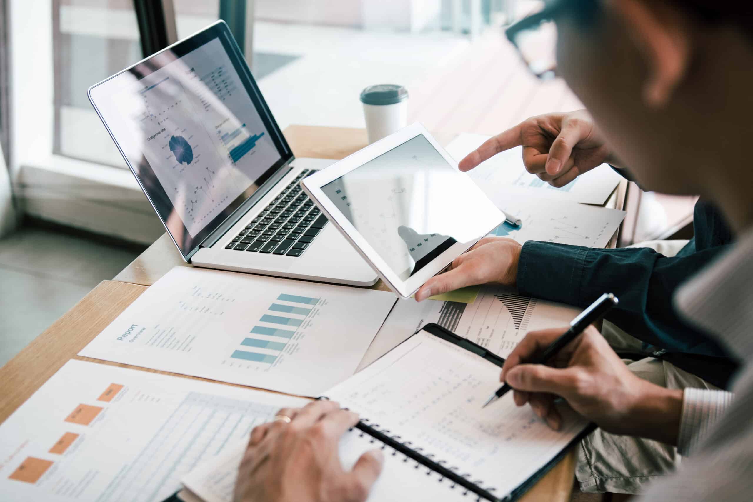 Two people review financial charts on paper, a tablet, and a laptop at a desk. One person points at the tablet whilst the other takes notes in a notebook. A coffee cup sits in the background.