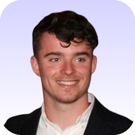 A young man with short brown hair wearing a dark jacket over a white shirt, smiling against a light purple gradient background.