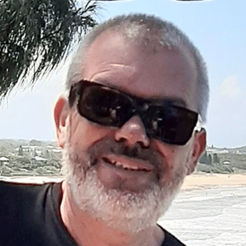 A smiling man with a grey beard and short hair wears dark sunglasses and a black shirt, standing outdoors near a beach with trees and blue sky in the background.