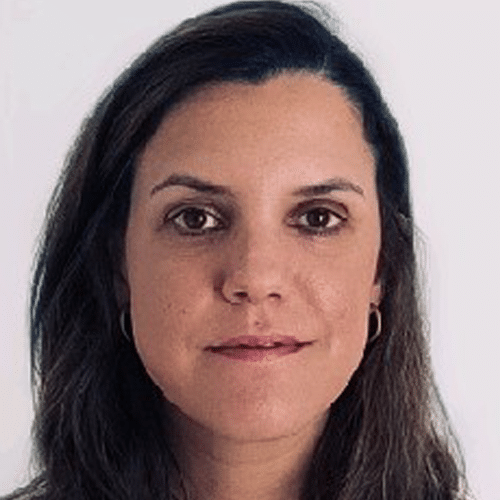 A woman with long dark hair, brown eyes, and light skin looks directly at the camera, wearing small hoop earrings. The background is plain and light-coloured.