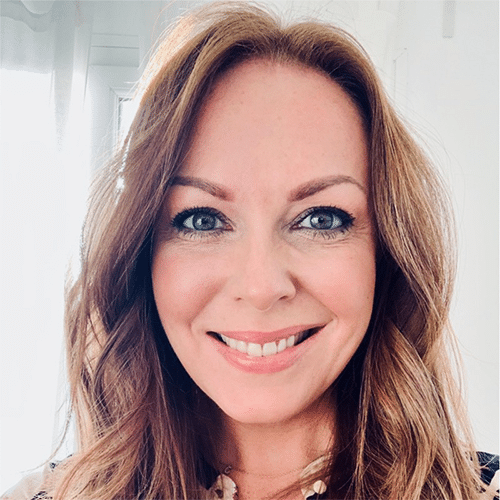 A woman with long, wavy light brown hair smiles at the camera. She has blue eyes and is standing in a brightly lit room with white walls and sheer curtains.