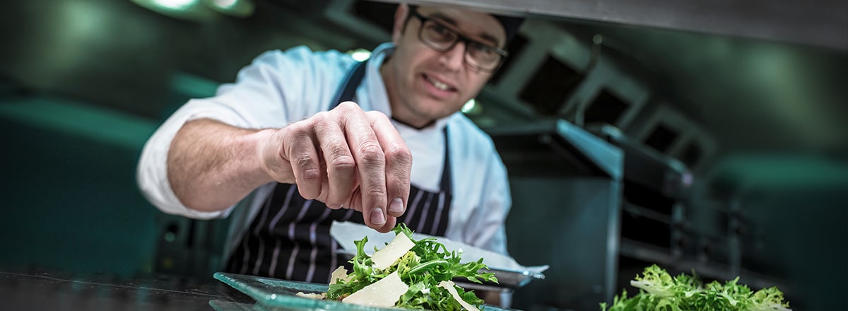 A chef in glasses and a striped apron is placing garnish on a plate of salad leaves in a restaurant kitchen.