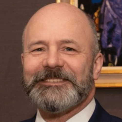 A middle-aged man with a bald head and grey beard smiles, wearing a suit. A colourful painting is visible in the background.