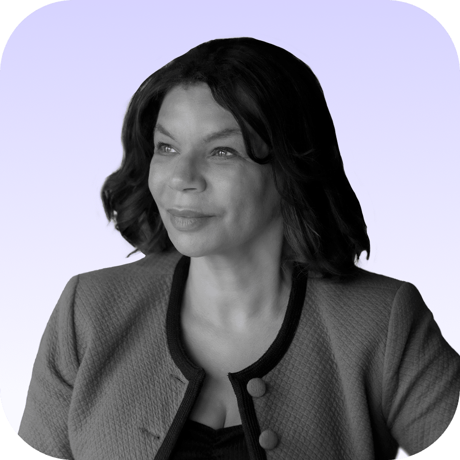 A woman with wavy dark hair, wearing a textured jacket over a top, looks slightly to the side and smiles. The background is a soft gradient from light purple to white, and the image is in black and white.