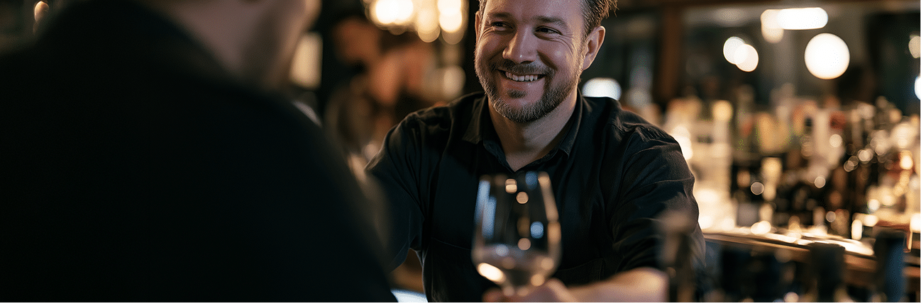 A smiling barman hands a glass of wine to a customer at a dimly lit bar, with bottles and warm lights visible in the background.