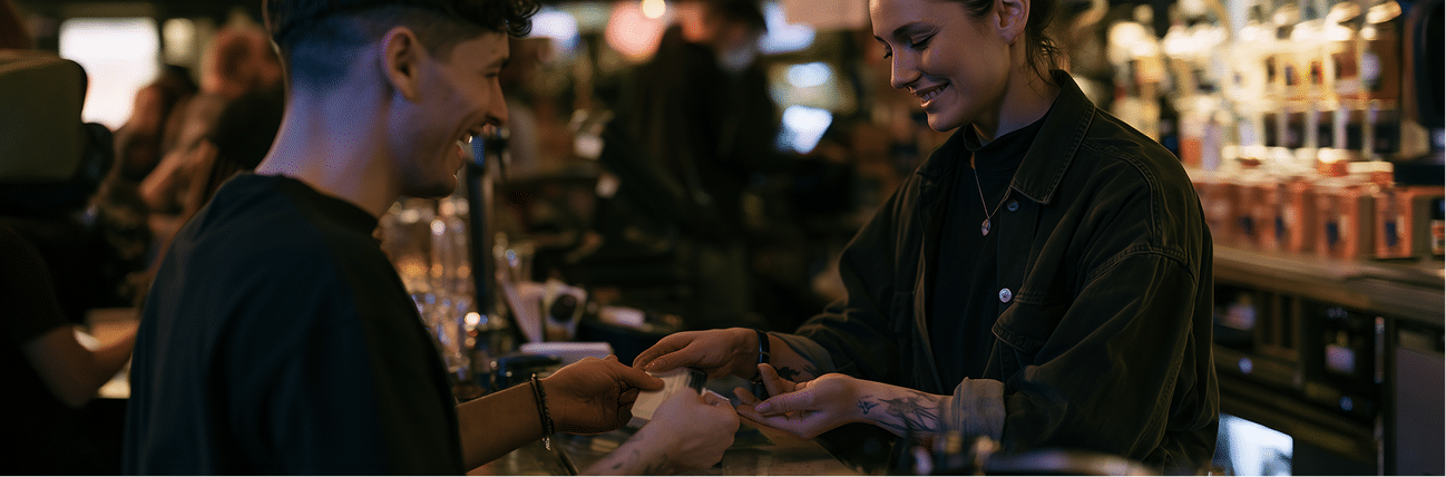A smiling barman hands a drink to a customer across a bar counter in a warmly lit, busy bar setting.