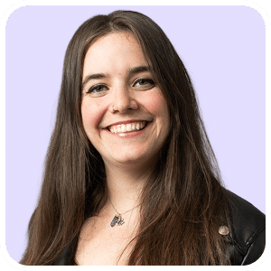 Smiling woman with long brown hair wearing a black jacket and necklace, standing in front of a light purple background.