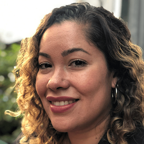 A woman with curly hair smiles at the camera outdoors. She is wearing hoop earrings and has a natural make-up look. The background is softly blurred with hints of greenery.
