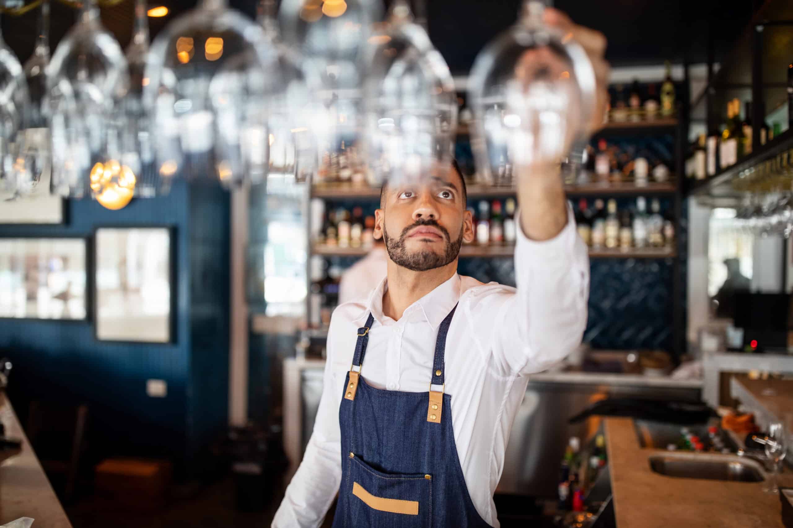 A barman in a white shirt and denim apron reaches up to grab a wine glass from a rack above a bar, with shelves of bottles in the background.