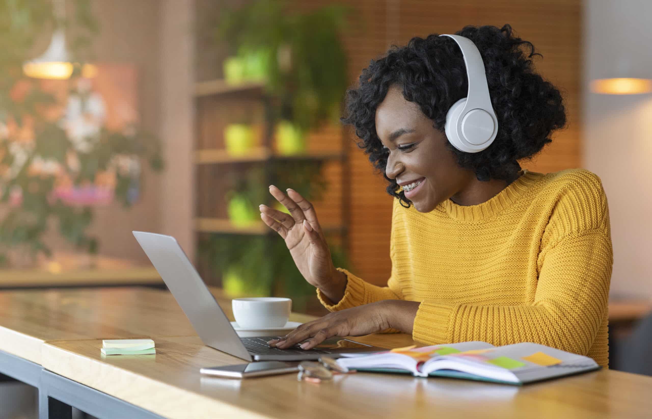 Woman in a yellow jumper wearing headphones smiles and waves at her laptop during a video call, sitting at a desk with a notebook, pen, and coffee cup in a cosy, well-lit room with plants in the background.