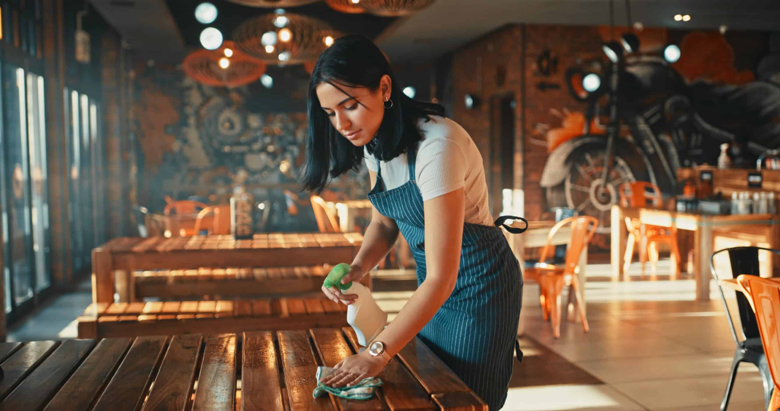 A woman in an apron cleans a wooden table in a stylish, empty restaurant with warm lighting and mural art on the brick walls.