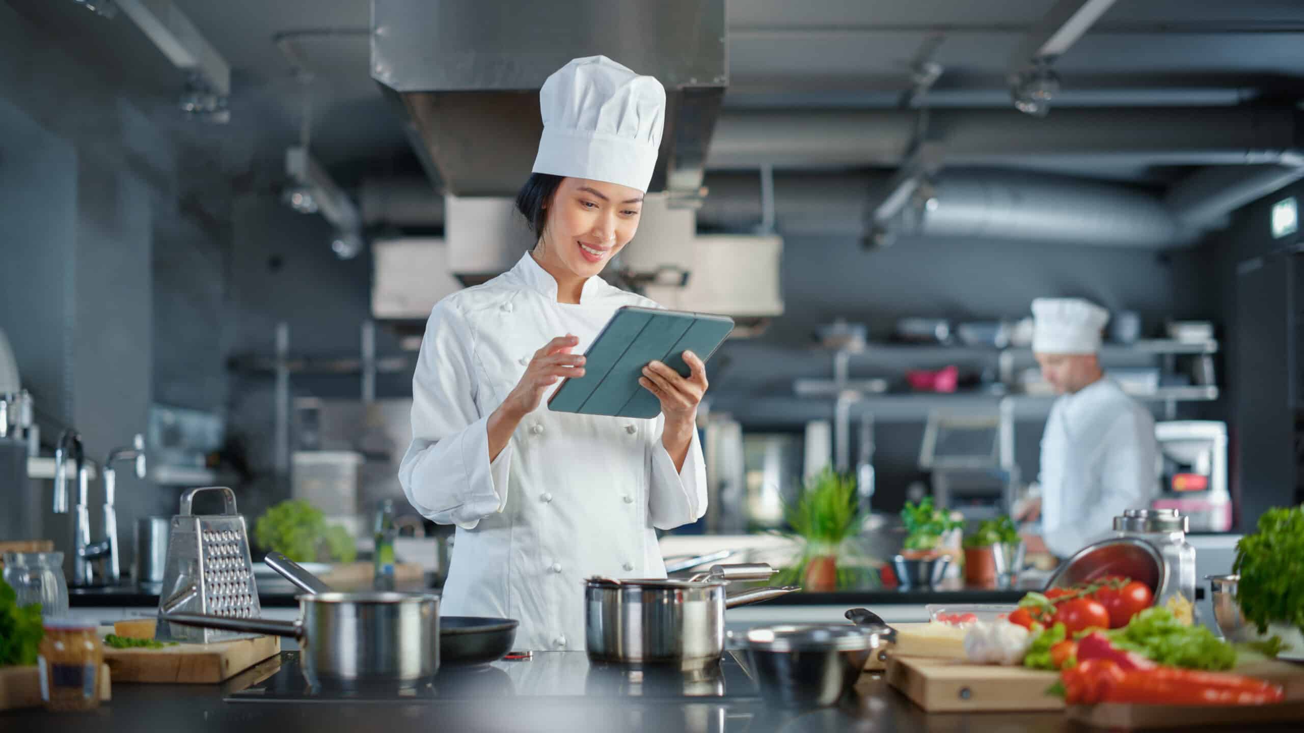 A chef in a white uniform and hat smiles whilst using a tablet in a modern kitchen. Stainless steel saucepans and chopped vegetables are on the worktop. Another chef works in the background.