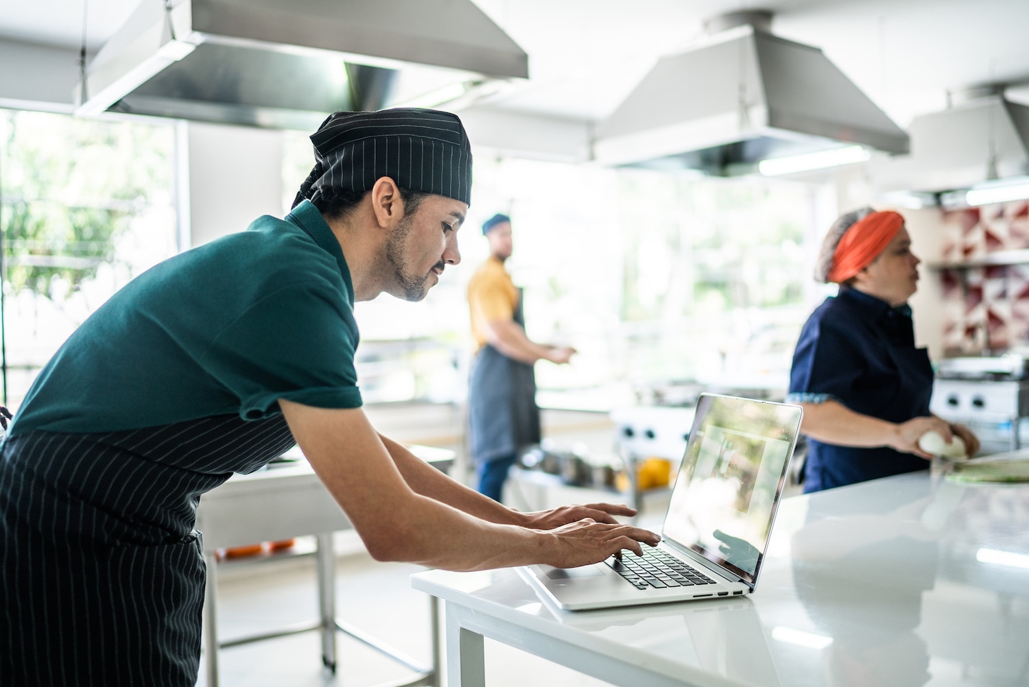 A chef in a striped apron and black hat types on a laptop in a bright, modern kitchen, whilst two other people work in the background.