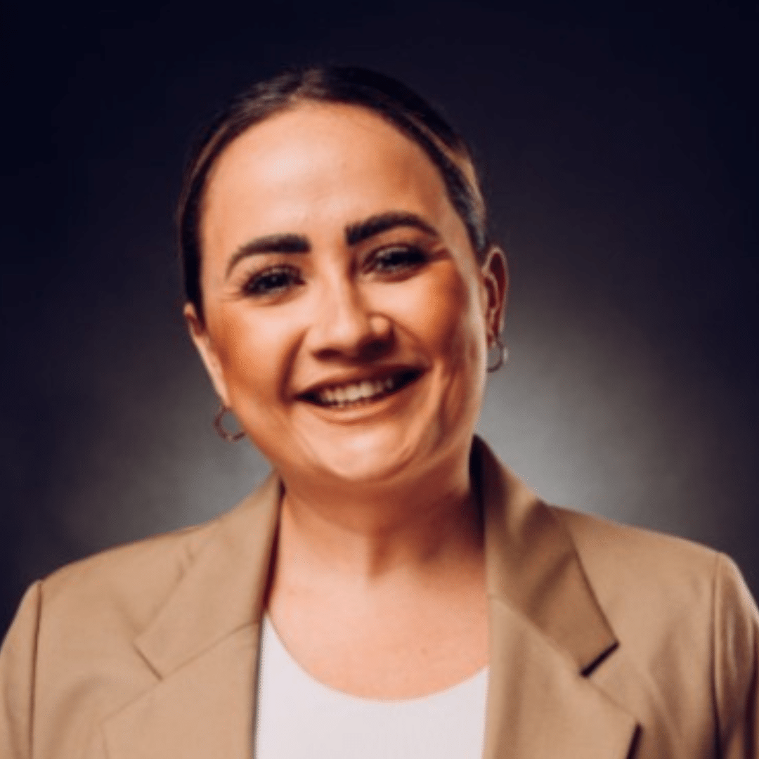 A woman with dark hair pulled back, wearing a tan blazer and white top, smiles at the camera against a dark, softly lit background.